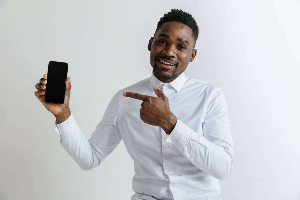 Retrato interior de un atractivo joven africano negro aislado sobre fondo rosa, sosteniendo un smartphone en blanco, sonriendo a la cámara, mostrando la pantalla, sintiéndose feliz y sorprendido. Emociones humanas, faciales
 - Foto, Imagen