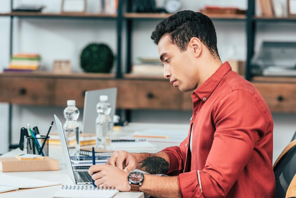 Concentrated student with tattoo in red shirt using laptop at desk - Photo, Image