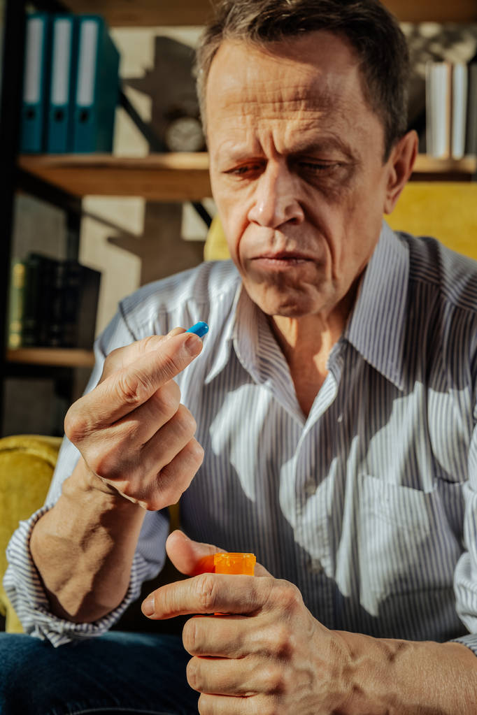 Сфокусированный старый расстроенный мужчина держит оранжевый контейнер с таблетками
 - Фото, изображение