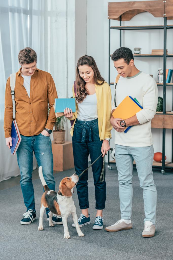 Full length view of three multiethnic smiling students with dog holding notebooks - Photo, Image