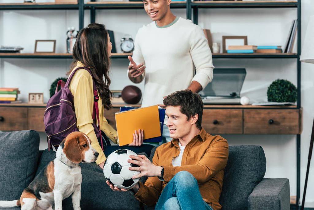 Estudantes multiétnicos com cadernos e bola de futebol sentados no sofá com cão
 - Foto, Imagem