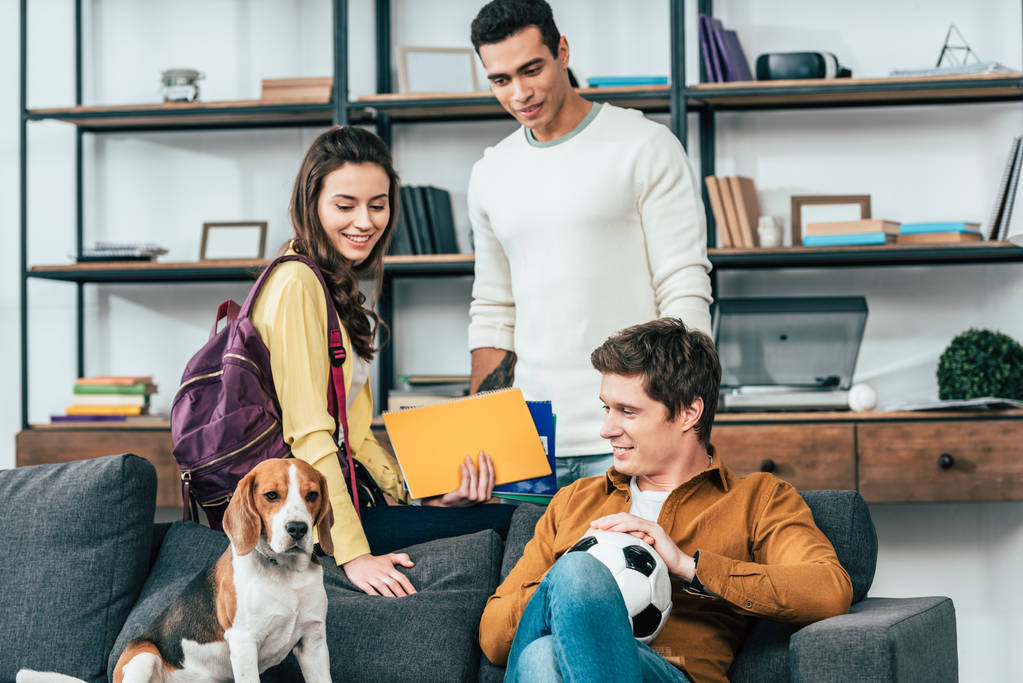 drei multikulturelle Studenten mit Notizbüchern und Fußballball auf Sofa mit Hund - Foto, Bild