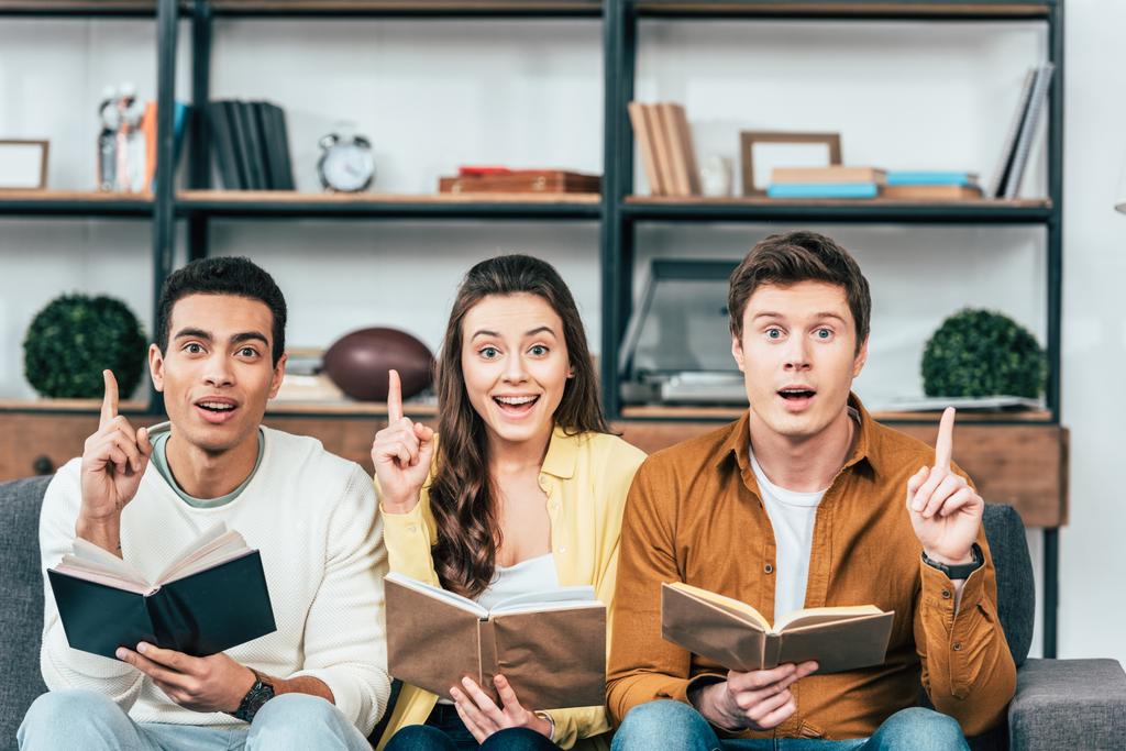 Three multiethnic students with books pointing with fingers up in living room - Photo, Image