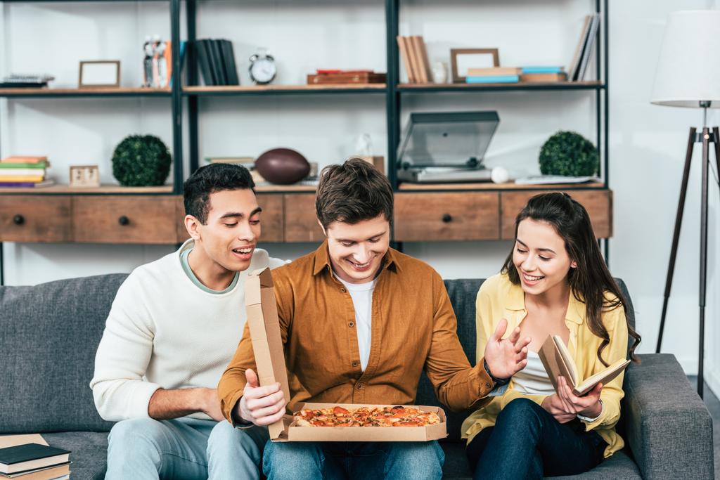 Трое смеющихся мультикультурных друзей сидят на диване с пиццей в гостиной
 - Фото, изображение