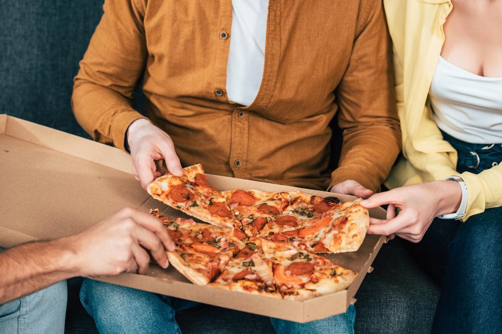 Oříznutý pohled na tři veselé přátele, kteří sedí na pohovce a jedí pizzu společně - Fotografie, Obrázek