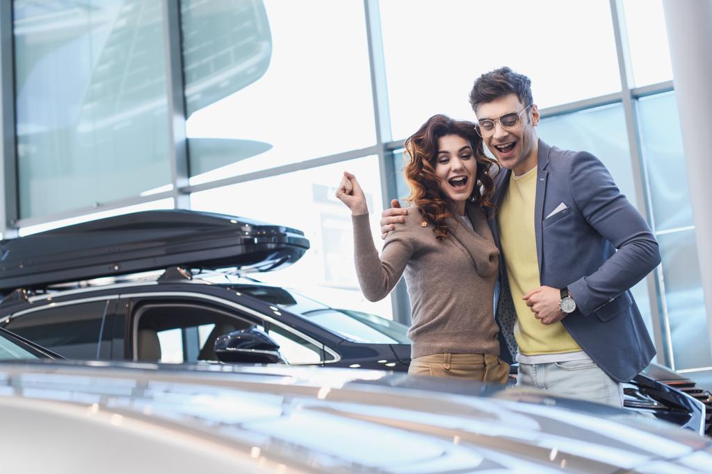 homem alegre abraçando encaracolado mulher atraente enquanto celebra triunfo no showroom carro
  - Foto, Imagem