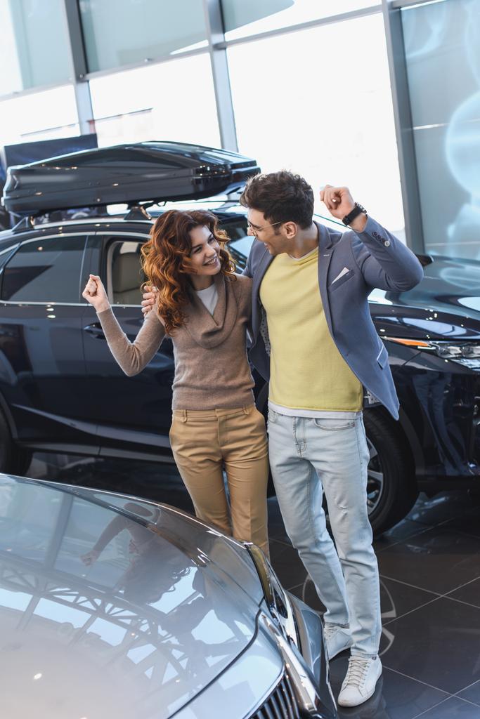 cheerful man in glasses hugging curly attractive woman while celebrating triumph in car showroom  - Photo, Image