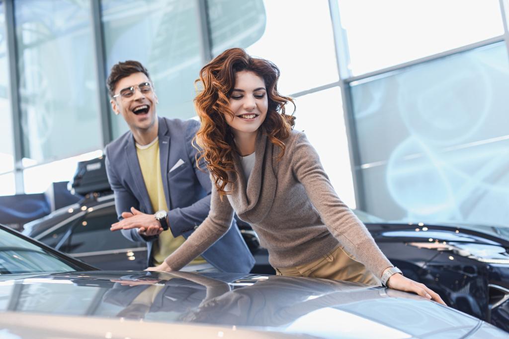 επιλεκτική εστίαση της χαρούμενη σγουρά γυναίκα χαμογελά κοντά ενθουσιασμένος άνθρωπος σε γυαλιά στην έκθεση αυτοκινήτων - Φωτογραφία, εικόνα