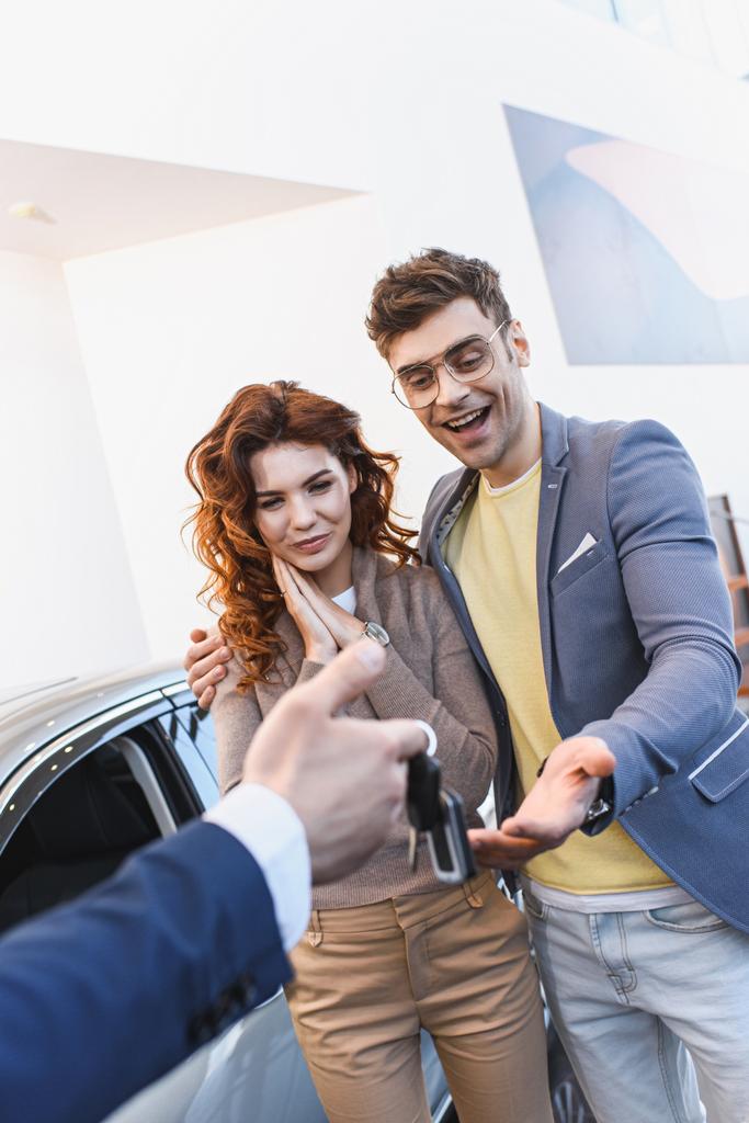 επιλεκτική εστίαση του ευτυχισμένο άνθρωπο και γυναίκα που αναζητούν τα κλειδιά στο χέρι του εμπόρου αυτοκινήτων στην έκθεση αυτοκινήτων  - Φωτογραφία, εικόνα