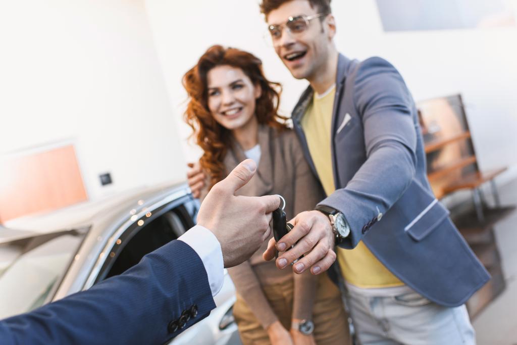 περικομμένη θέα του εμπόρου αυτοκινήτων δίνοντας κλειδιά για χαρούμενο άντρα και γυναίκα σε εκθεσιακός χώρος  - Φωτογραφία, εικόνα