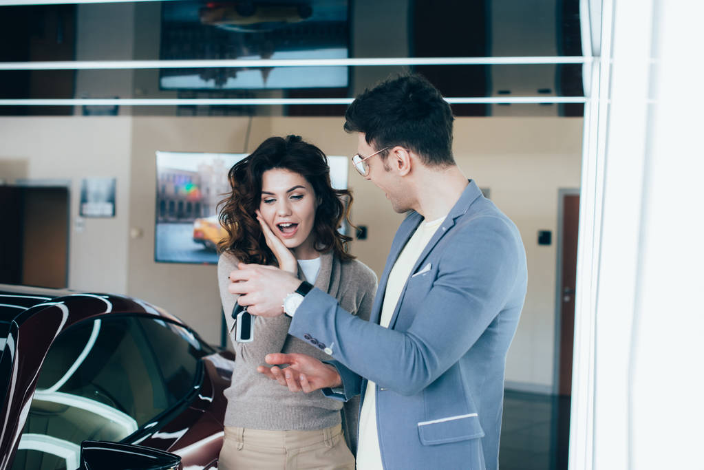 cheerful man holding keys near surprised curly girl while standing near red automobile - Photo, Image
