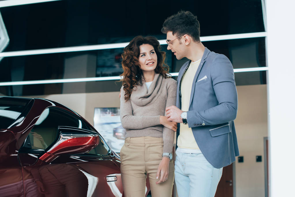 счастливый мужчина смотрит на кудрявую девушку, стоя возле красного автомобиля
 - Фото, изображение