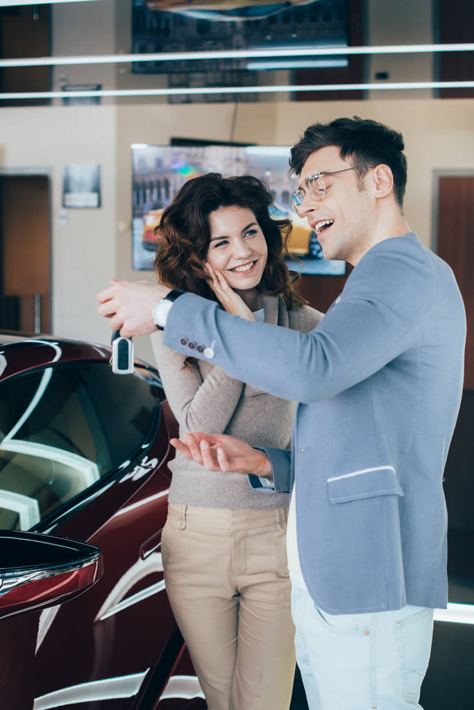 szczęśliwy kobieta patrząc na przystojny mężczyzna trzymając klucze w pobliżu czerwony samochód - Zdjęcie, obraz