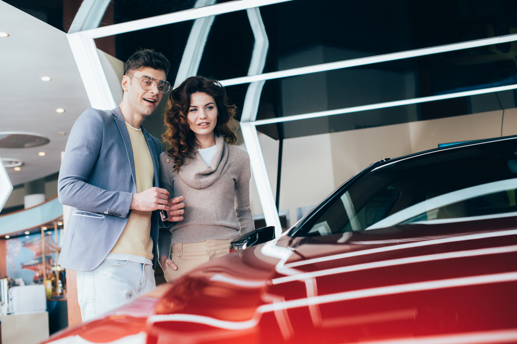веселый человек держит ключи, стоя с кудрявой девушкой рядом с красным автомобилем
 - Фото, изображение