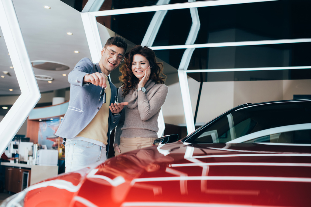 homem alegre em óculos segurando chaves enquanto está com a menina sorridente perto do automóvel vermelho
 - Foto, Imagem