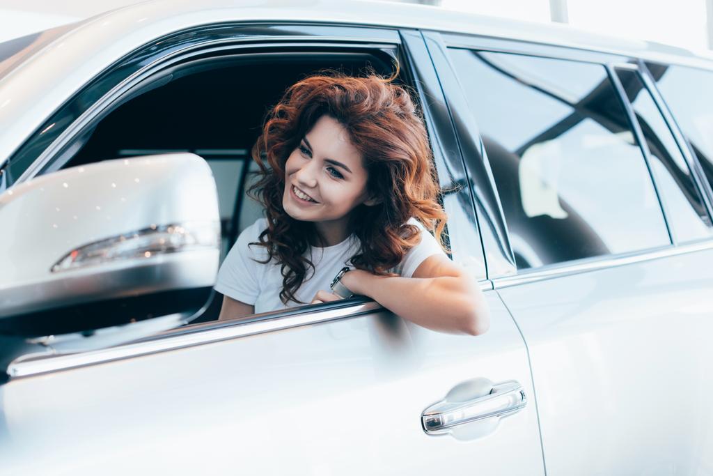selectieve focus van vrolijke krullende vrouw zitten en glimlachen in de auto  - Foto, afbeelding