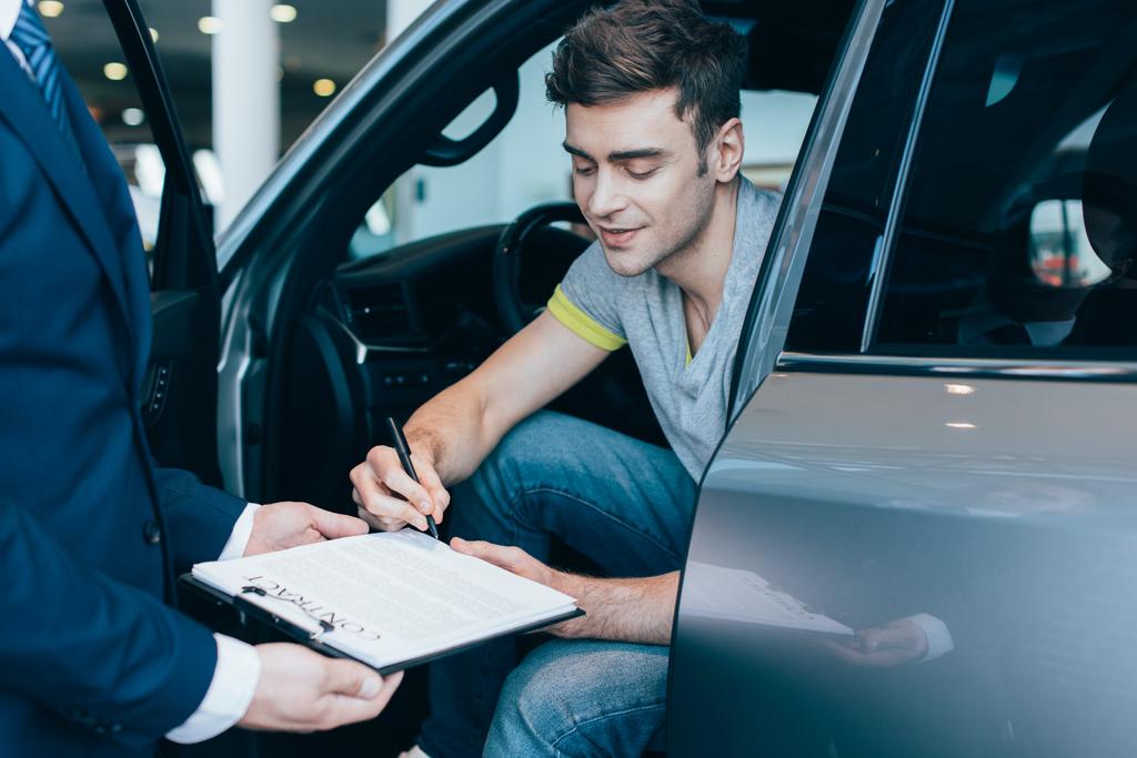 περικομμένη προβολή του εμπόρου αυτοκινήτου κρατώντας πρόχειρο, ενώ χαρούμενο άντρα υπογραφή σύμβαση - Φωτογραφία, εικόνα