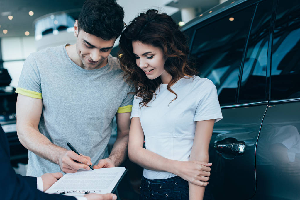 私は幸せな男は魅力的なカーリーの女性の近くに契約を署名しながらクリップボードを保持する車のディーラーのクロップビュー   - 写真・画像