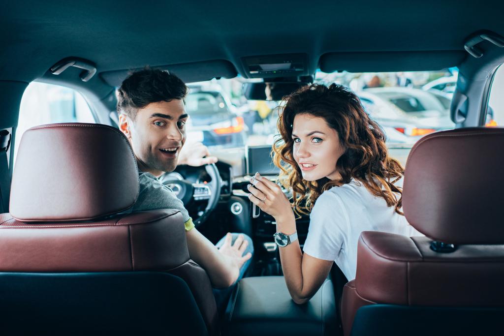 επιλεκτική εστίαση του ευτυχισμένο άνθρωπο και σγουρά χαρούμενη γυναίκα κοιτάζοντας την κάμερα, ενώ κάθονται στο αυτοκίνητο  - Φωτογραφία, εικόνα