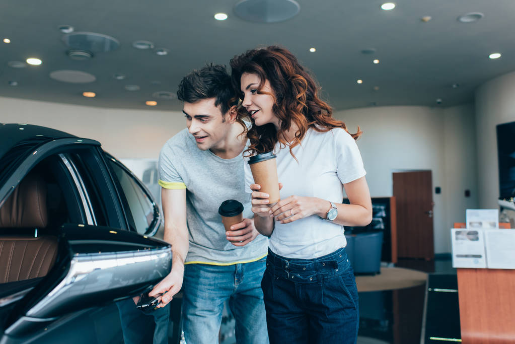 cheerful man holding paper cup near curly girl while looking at automobile  - Photo, Image