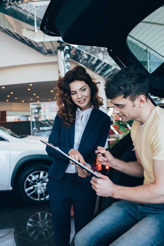 Happy man Signing paper in de buurt van glimlachende auto dealer in auto showroom - Foto, afbeelding