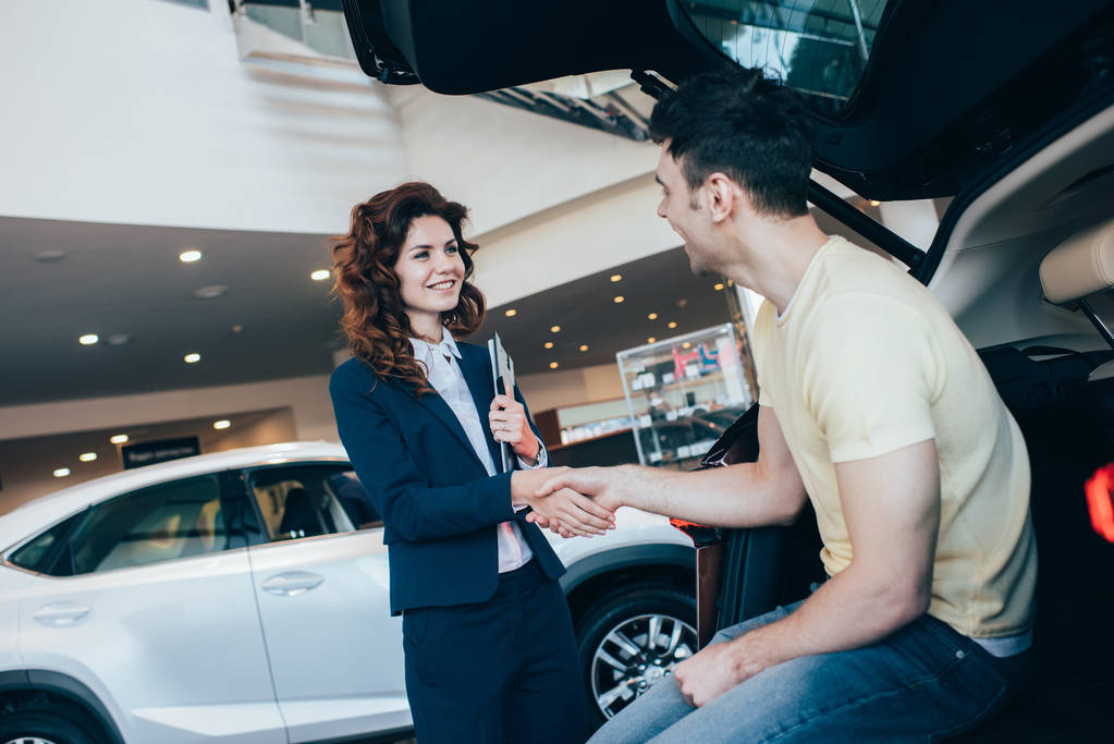 vrolijke auto dealer en klant schudden handen terwijl staande in de buurt van nieuwe auto - Foto, afbeelding