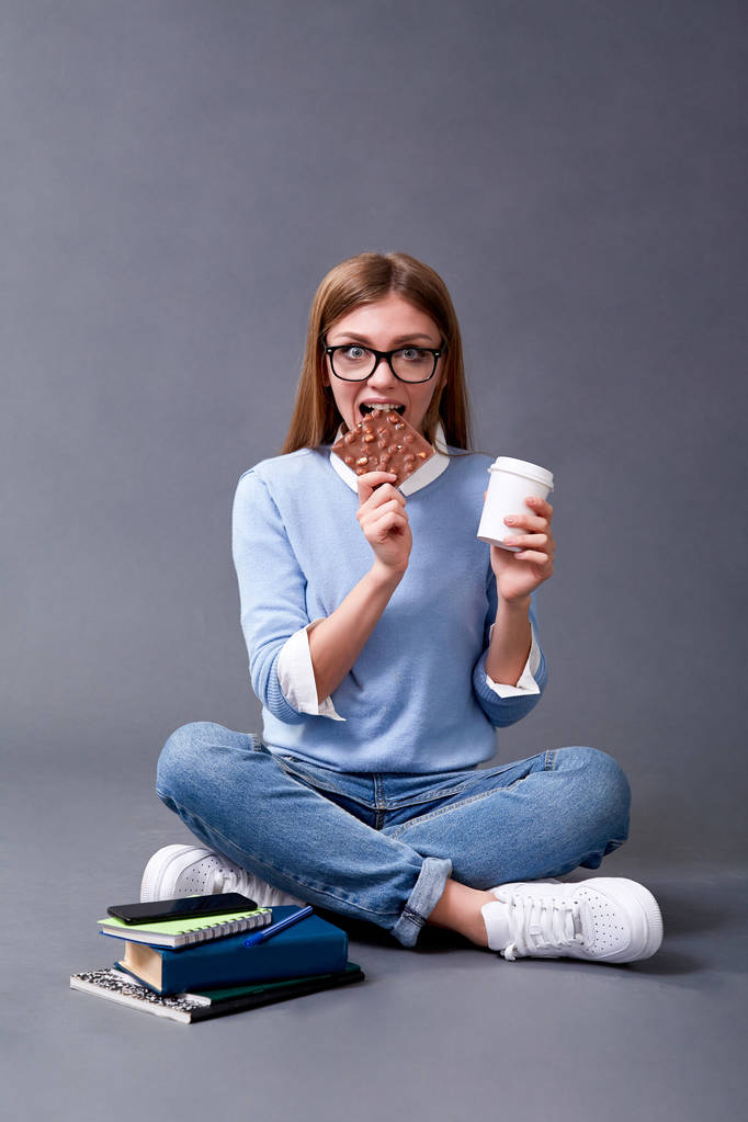Uno studente mangia una barretta di cioccolato, beve caffè e sorride. Concetto studentesco. Studio.
 - Foto, immagini