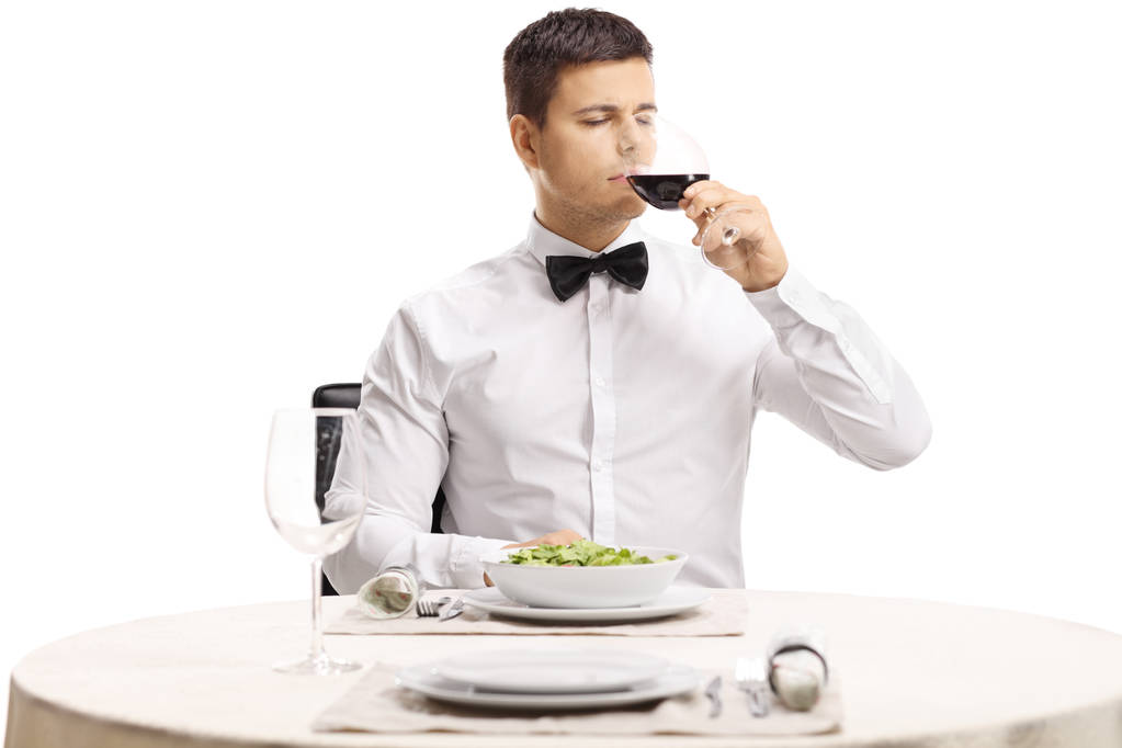 Portrait of a young handsome man with a bow tie tasting red wine at a restaurant table isolated on white background - Photo, Image