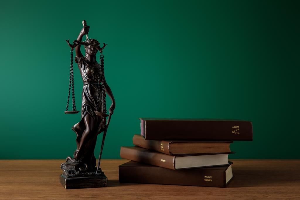 бронзовая статуэтка с весами справедливости и объемами книг на деревянном столе на темно-зеленом фоне
 - Фото, изображение