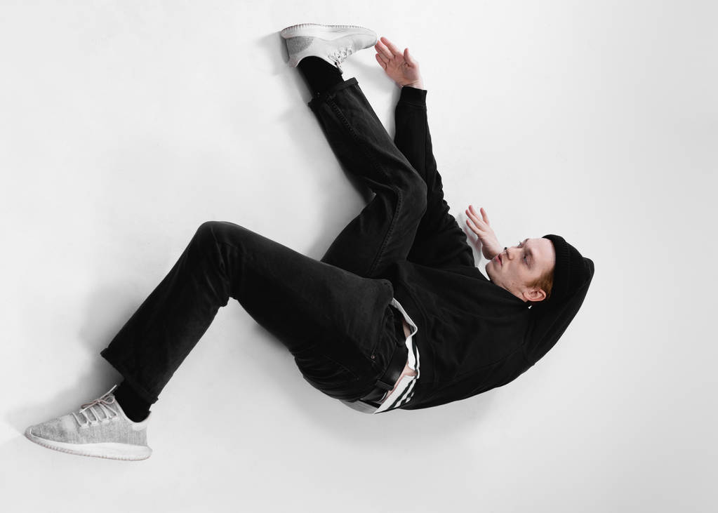 Freestyle danser gekleed in zwarte jeans, trui, hoed en grijze sneakers is dansen liggend op de vloer in de studio op de witte achtergrond - Foto, afbeelding