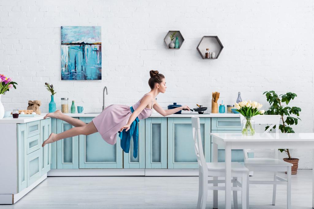 mooi barefoot meisje in elegante jurk en schort vliegen in de lucht met de pan in de keuken - Foto, afbeelding