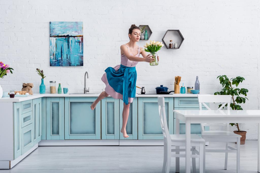 mooi barefoot meisje in elegante jurk en schort vliegen in de lucht met boeket tulpen in glazen pot in de keuken - Foto, afbeelding