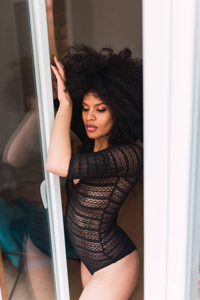 Nuori viehättävä musta nainen kehonpuku poseeraa lähellä ikkunaa
 - Valokuva, kuva