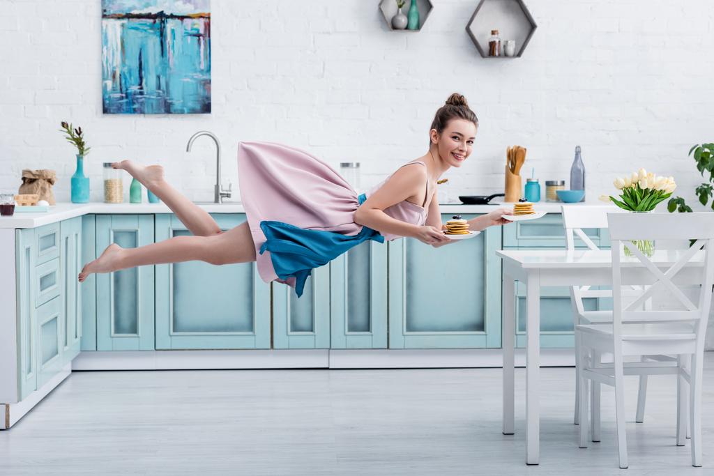 junge lächelnde Barfußfrau schwebt in der Luft, während sie leckere Pfannkuchen serviert - Foto, Bild