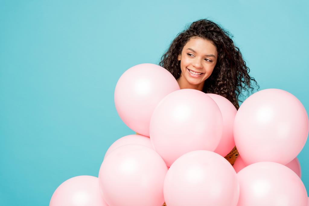 Pretty Curly Afro-Amerikaanse meisje glimlachen in de buurt van roze lucht ballonnen geïsoleerd op blauw  - Foto, afbeelding