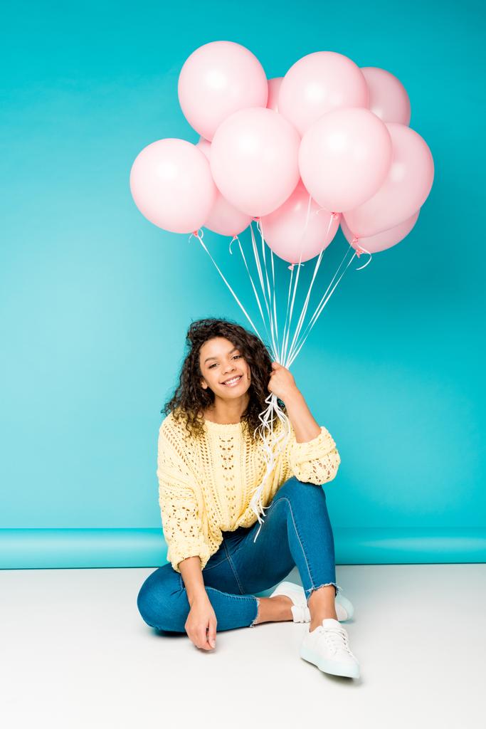 Pretty Curly Afro-Amerikaanse meisje zitten met roze lucht ballonnen op blauw  - Foto, afbeelding