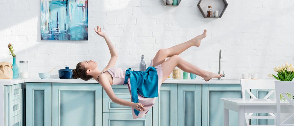 plano panorámico de mujer joven en delantal y vestido levitando en el aire en la cocina
 - Foto, imagen