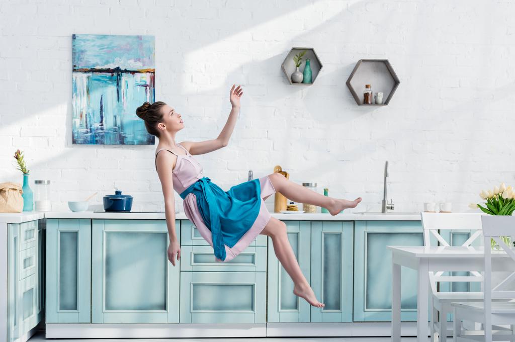  young elegant woman in apron and dress flying in air in kitchen - Photo, Image