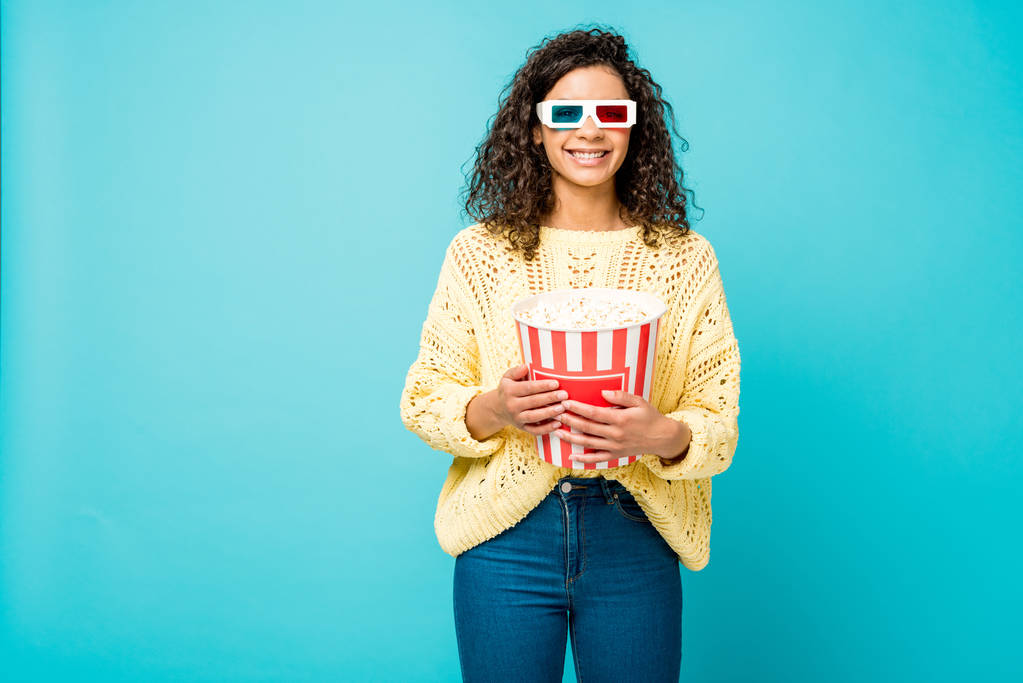 Wesoła kręcone African American kobieta trzyma wiadro popcorn i uśmiechając się w okulary 3D na niebiesko  - Zdjęcie, obraz