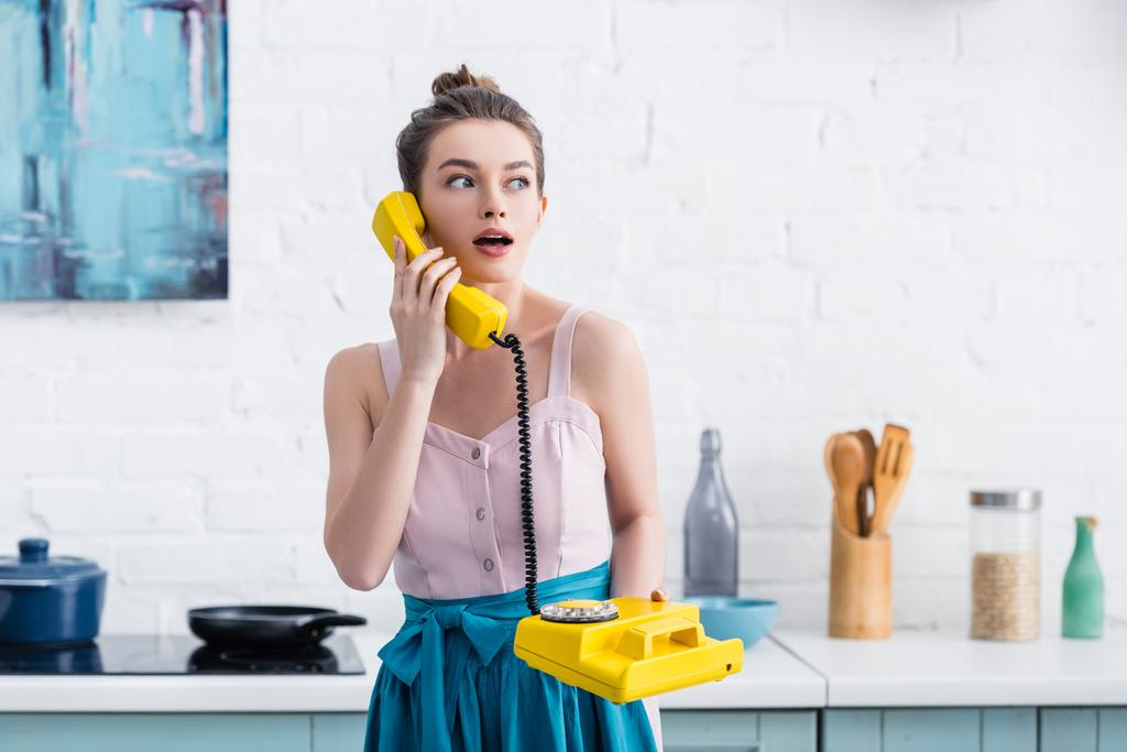 σοκαρισμένος γυναίκα μιλάμε για ρετρό κίτρινου τηλεφωνικό στην κουζίνα   - Φωτογραφία, εικόνα