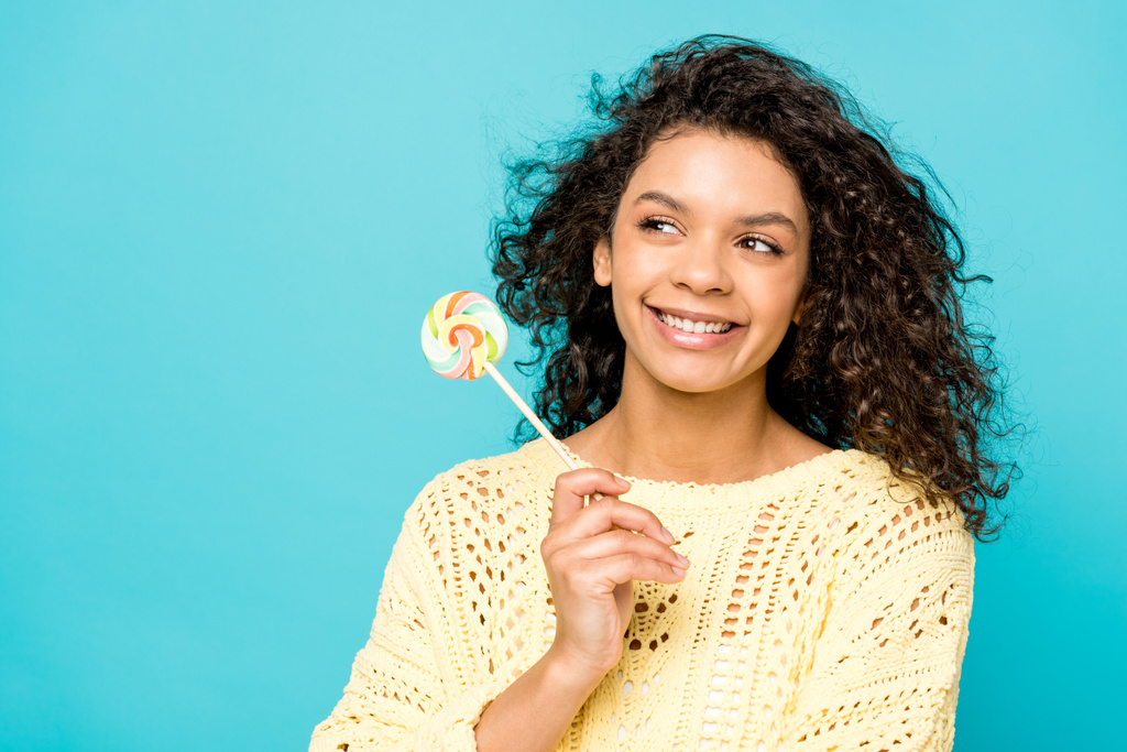 happy curly african american girl smiling while holding lollipop isolated on blue - Photo, Image