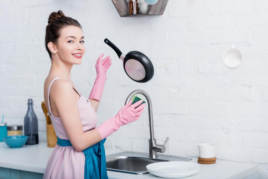 χαμογελά ευτυχισμένη νεαρή γυναίκα στο λαστιχένια γάντια βλέπουν κάμερα ενώ μαγειρικά σκεύη αιωρούμενο στον αέρα  - Φωτογραφία, εικόνα