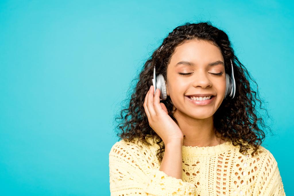 счастливая кудрявая африканская американская девушка слушает музыку в наушниках с закрытыми глазами, изолированными на голубом
 - Фото, изображение