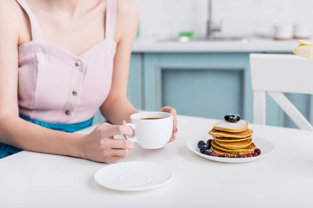 обрезанный вид молодой женщины, держащей чашку кофе возле тарелки с блинами на столе
 - Фото, изображение