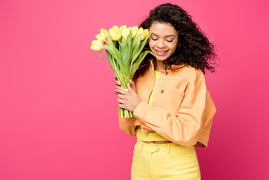 привлекательная улыбающаяся африканская американка, держащая желтые тюльпаны изолированные на малиновом
 - Фото, изображение