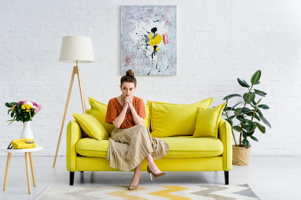 nachdenklich elegante junge Frau sitzt mit gekreuzten Beinen und geballten Händen auf gelbem Sofa im Wohnzimmer - Foto, Bild