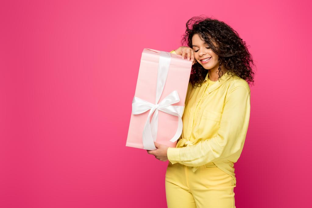 glücklich lockige afrikanisch-amerikanische Frau hält rosa Geschenkbox mit weißem Satinband isoliert auf purpurrot  - Foto, Bild