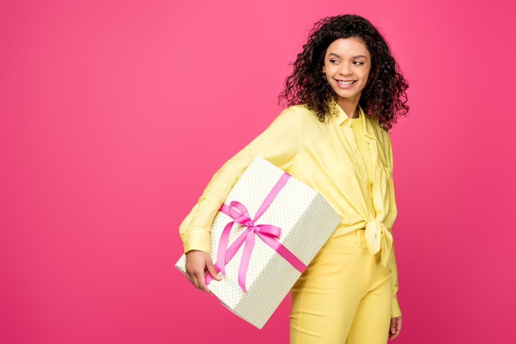 fröhlich lockige afrikanisch-amerikanische Frau hält Geschenkbox mit rosa Satinband isoliert auf purpurrot  - Foto, Bild