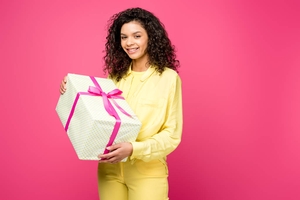 Happy Curly Afro-Amerikaanse vrouw Holding gift box met roze satijnen lint geïsoleerd op Crimson  - Foto, afbeelding
