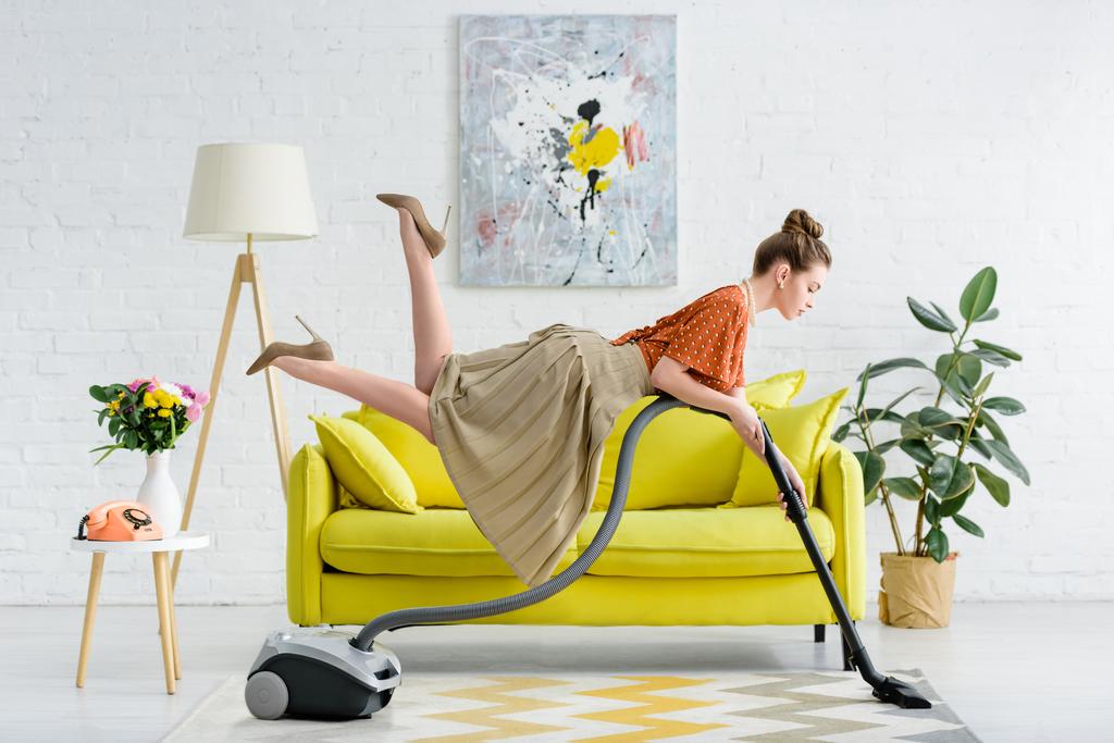 vista lateral de elegante mujer joven concentrada levitando en el aire y la alfombra de aspiración en la sala de estar
 - Foto, Imagen
